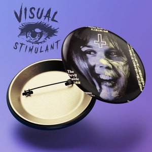 May include: A black and white button featuring a close-up of a woman's face with a cross over her forehead. The text "The Devil Inside" is printed on the button. The button is attached to a gold-colored pin back.