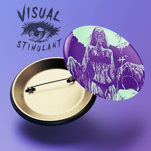 May include: A purple and green button with a graphic of a woman with long hair and a creepy expression. The text "サザザ" is printed on the button. The back of the button is gold.