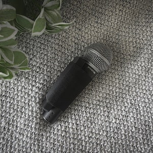 May include: A black microphone with a silver grill and a black foam cover. The microphone is labeled "Dynamic" and is lying on a gray knitted surface.