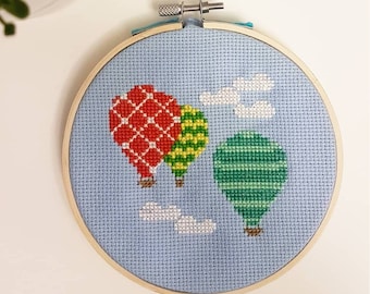 May include: A blue and white cross-stitch embroidery hoop with a design of three hot air balloons in red, green, and yellow with white clouds.