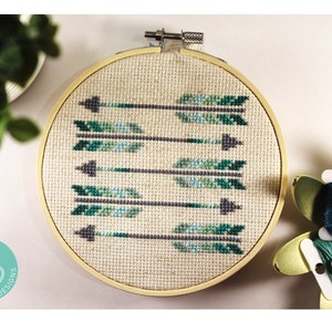 Puede incluir: Un aro de bordado de punto de cruz terminado con un diseño de seis flechas verdes y grises. El aro está sobre una superficie blanca con una planta verde y un carrete de hilo turquesa en el fondo.