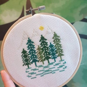 May include: A white embroidery hoop with a cross-stitch design of a forest scene. The design features five green trees with a yellow sun in the sky above them.