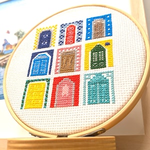 Peut inclure: Un cercle de broderie rond avec un fond en tissu blanc et des motifs de point de croix colorés de neuf portes différentes. Les portes sont de différentes couleurs, notamment le bleu, le jaune, le rouge, le vert et le brun. Le cercle est sur un support en bois.