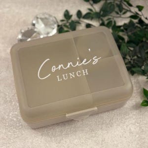 May include: A beige rectangular lunch box with the text "Connie's LUNCH" in white script. The lunch box has a snap closure and is divided into sections. The background includes greenery and a decorative crystal.