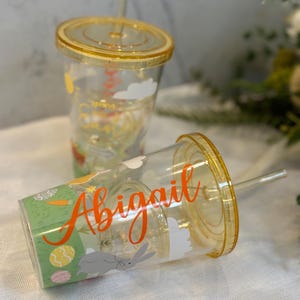 May include: Two clear plastic tumblers with yellow lids and straws. One is upright, the other tilted. The tumblers feature a spring design with a bunny, eggs, and the name "Abigail" in orange script.