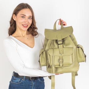 Puede incluir: Una mochila de cuero verde claro con varios bolsillos y cierre de hebilla. La mochila tiene correas ajustables y un asa superior.