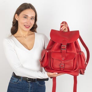 May include: A red leather backpack with multiple pockets and a buckle closure. The backpack has a top flap with a buckle closure and adjustable straps.