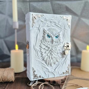 Op de afbeelding: Wit leren dagboek met een reliëf van een uil en een zilveren slot en sleutel. De uil heeft blauwe ogen en is omgeven door een zilveren frame.