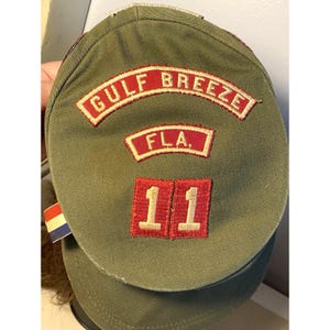 May include: Green canvas scout hat with red and white embroidered patches. The patches read "GULF BREEZE FLA." and "11".