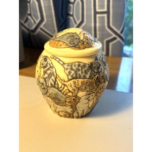 May include: A cream-colored ceramic jar with a matching lid, featuring a detailed relief design of multiple cats in various poses. The cats are rendered in shades of gray, brown, and tan, covering the entire exterior surface of the vessel.