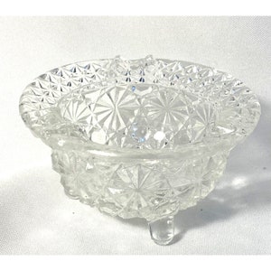 May include: A clear glass bowl with a starburst pattern and three legs. The bowl is made of pressed glass and has a vintage look.