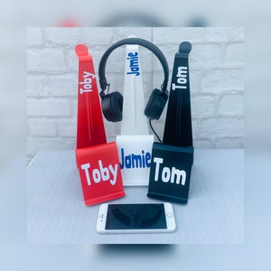 May include: Three headphone stands with personalised names: Toby, Jamie, and Tom. The stands are red, white, and black. A black smartphone is in the foreground.