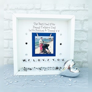 May include: White picture frame with a photo of a father and two children on a beach. The frame has a blue background and the words "The Best Dad Ever, Happy Father's Day, Love Hannah & Tommy x x" are written on it. The words "WE LOVE YOU" are spelled out with white tiles and a silver heart is in the bottom right corner.