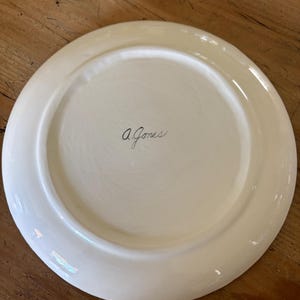 May include: A cream-colored ceramic plate with a wide rim. The underside of the plate features the handwritten signature "A. Jones" in black ink. The plate is set against a wooden surface.
