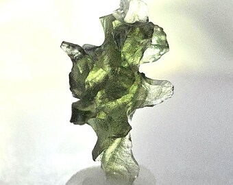May include: A translucent, olive-green moldavite gemstone with an irregular, textured surface. The natural glass formation has a unique, organic shape, and is displayed against a white background. The gemstone is a striking example of natural crystal formations.