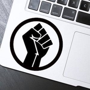 May include: Black and white sticker of a raised fist in a circle on a white laptop.