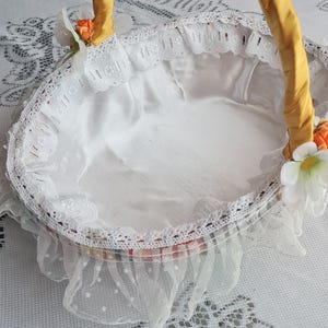 以下が含まれることがあります： 白いバスケットに黄色のサテンのハンドルと白いレースのトリムが付いています。バスケットは白いサテンで裏打ちされており、側面に小さなオレンジの花が付いています。