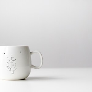 Pu&ograve; includere: Una tazza in ceramica bianca con un design geometrico in bianco e nero di una costellazione sul lato.