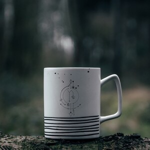 Pu&ograve; includere: Tazza in ceramica bianca con un design geometrico in bianco e nero con frecce, cerchi e stelle. La tazza &egrave; appoggiata su un tronco d'albero.