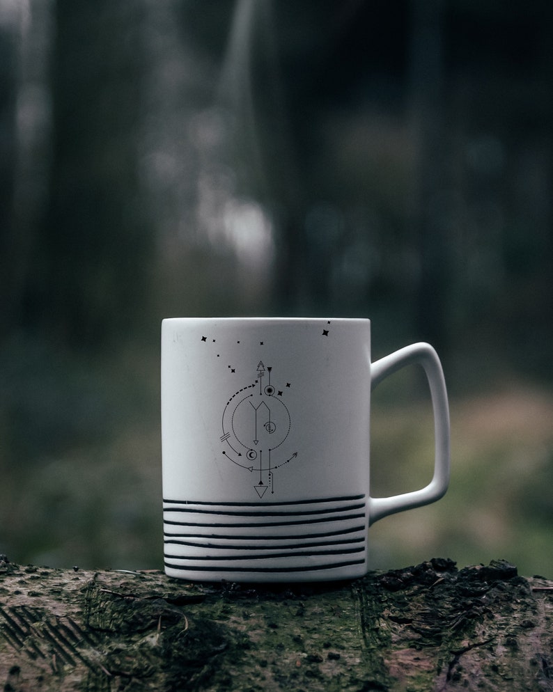 Pu&ograve; includere: Tazza in ceramica bianca con un design geometrico in bianco e nero con frecce, cerchi e stelle. La tazza &egrave; appoggiata su un tronco d'albero.