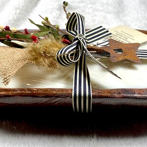 Puede incluir: Un bol de madera para masa relleno con una vela blanca y envuelto con una cinta a rayas blancas y negras. El bol está decorado con una estrella de arpillera, flores secas y una estrella de metal oxidado.