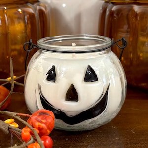 Peut inclure: Une bougie en pot de verre transparent remplie de cire blanche, arborant un motif de visage de citrouille d'Halloween noir sur le devant. Le pot est doté d'une anse en fil de fer noir fixée au bord. La bougie est entourée de petites baies décoratives orange.