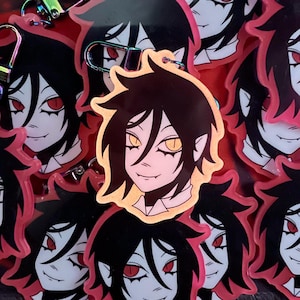 May include: A collection of anime-style keychains featuring a character with black hair, red eyes, and a pale complexion. The keychains have a gold border and a metal clasp. The background is a deep red.
