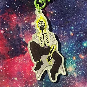 May include: A clear acrylic keychain featuring a seated skeleton character with a yellow crown and accents. The skeleton wears a white lab coat, black pants, and black shoes. The keychain has a green clasp and a galaxy background.