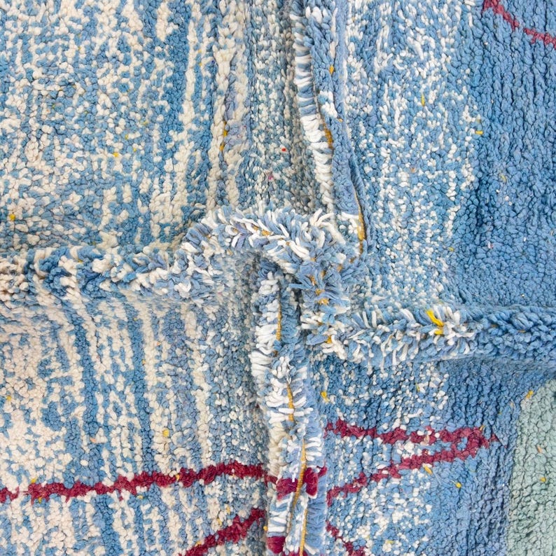 May include: Close-up of a textured rug with a blue and white color scheme. The rug features a mix of light and dark blue hues, with white accents and red stripes. The design includes a fringed border.