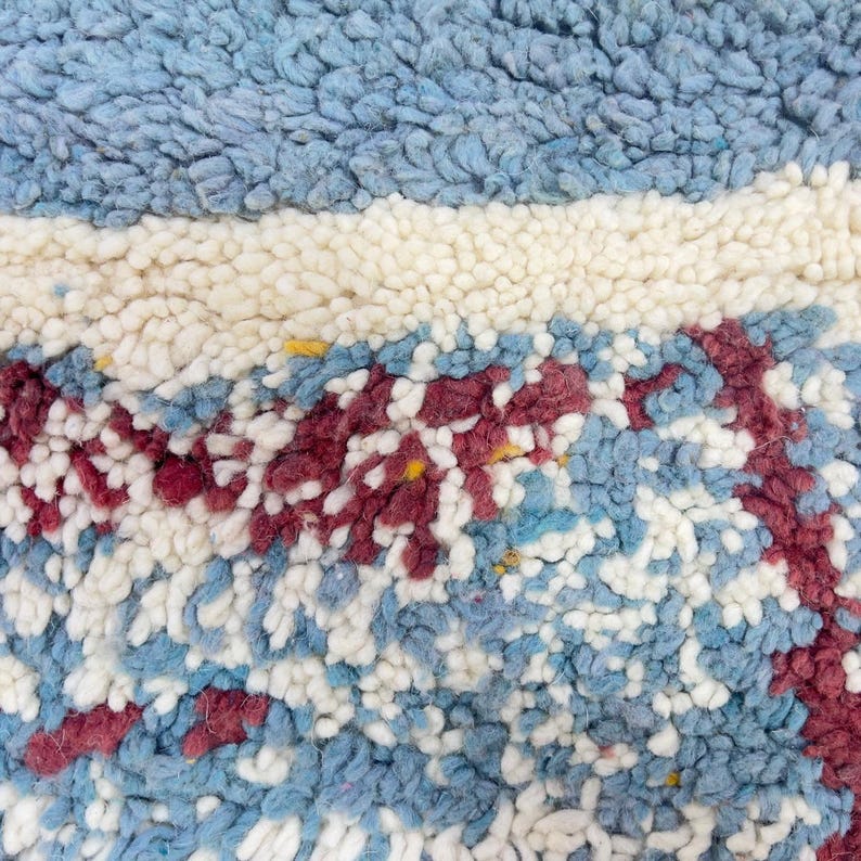 May include: Close-up of a textured rug with a mix of colors. The rug features a light blue top section, a white stripe, and a lower section with blue, white, and burgundy patterns. The rug has a soft, plush appearance.