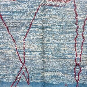 May include: Close-up of a blue and white rug with a textured surface. The rug features a geometric design with red lines. The rug's color palette includes shades of blue, white, and red. The rug is a decorative home decor item.
