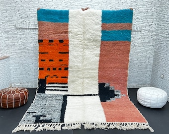 May include: A colorful Moroccan rug with geometric patterns in shades of orange, white, black, and blue. The rug has a fringe border and is made of wool.