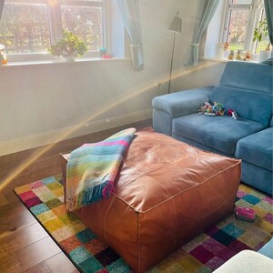 May include: A large, square, brown leather ottoman sits on a colorful rug. A multi-colored blanket with fringe is draped over the ottoman. A blue sectional sofa is in the background, along with a window with curtains.
