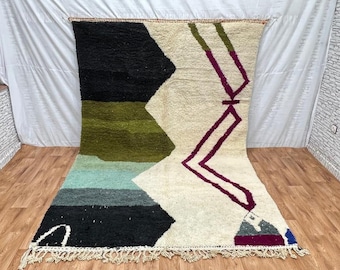 May include: A large, rectangular rug with a geometric design. The rug features a cream-colored background with black, green, blue, and purple shapes. The rug has a fringe along the bottom edge and is made of a thick, textured material.