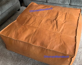 May include: A large, square ottoman in a warm, tan color. The leather surface is divided into rectangular sections by stitched seams. The ottoman is placed on a gray carpet, with a gray sofa visible in the background.