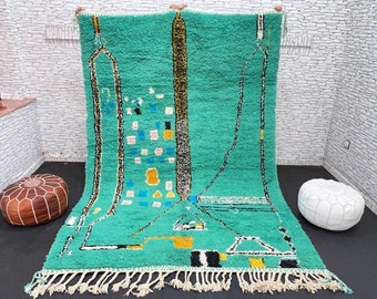 May include: A hand-woven Moroccan rug with a green background and geometric patterns in black, white, yellow, and blue. The rug has a fringe border.