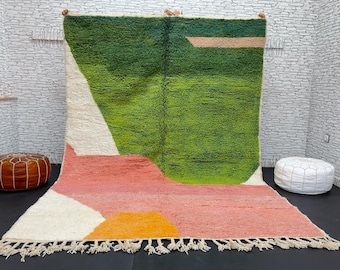 May include: A large, rectangular rug with an abstract design. The rug features a vibrant color palette, including green, pink, white, and yellow. The design incorporates geometric shapes and a fringed edge. The rug is displayed against a white brick wall.