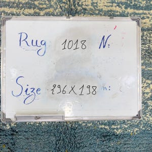 May include: A white board with the word "Rug" and the number "1018" written in blue ink. The word "Size" is also written in blue ink, followed by the dimensions "296 x 198". The rug is visible in the background.