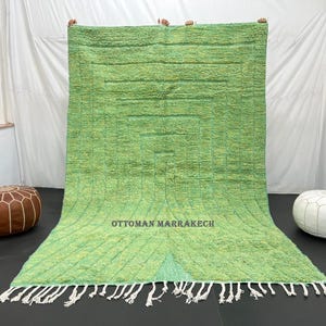 May include: A green and white Moroccan rug with a geometric pattern and fringe. The rug is laid out on a white floor with two poufs, one brown and one white.