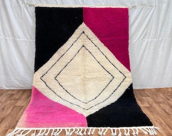 Könnte beinhalten: Ein weißer, schwarzer und rosa marokkanischer Teppich mit einem Diamantenmuster in der Mitte. Der Teppich hat einen Fransenrand.