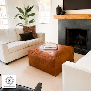 May include: A living room scene featuring a large, square, brown leather ottoman with books on top. Two white sofas and a fireplace with a wooden mantel are visible. A large potted plant adds a touch of greenery to the room. The room has a neutral color palette.