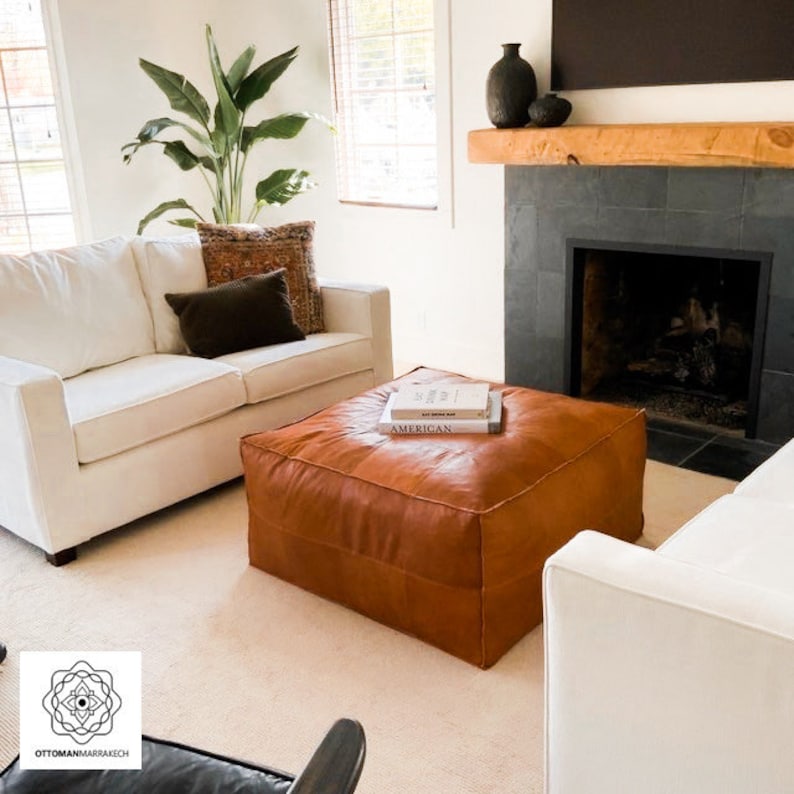May include: A living room scene featuring a large, square, brown leather ottoman with books on top. Two white sofas and a fireplace with a wooden mantel are visible. A large potted plant adds a touch of greenery to the room. The room has a neutral color palette.