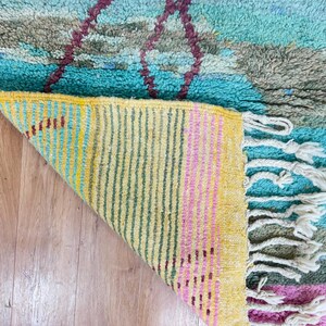May include: A colorful, handmade rug with a geometric design. The rug features a turquoise, gray, and burgundy pattern on the top, with a yellow, pink, and green striped underside. White fringe is visible on the edge.