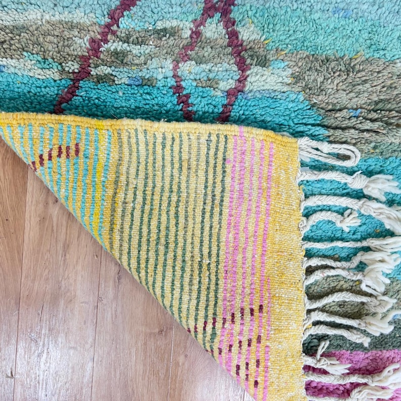 May include: A colorful, handmade rug with a geometric design. The rug features a turquoise, gray, and burgundy pattern on the top, with a yellow, pink, and green striped underside. White fringe is visible on the edge.