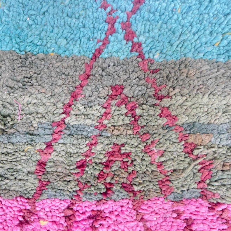 May include: Close-up of a textured rug with a geometric design. The rug features a color palette of blue, gray, and pink. A pink geometric pattern is overlaid on the gray section. The rug has a plush, soft appearance.