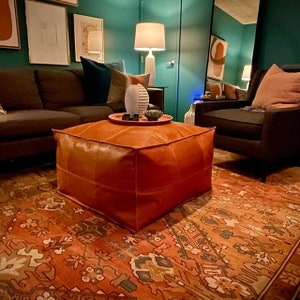 May include: A square, orange leather ottoman sits on a patterned rug in a living room setting. The ottoman is the focal point, with a tray and decorative items on top. A sofa and armchair are visible in the background, along with framed artwork.