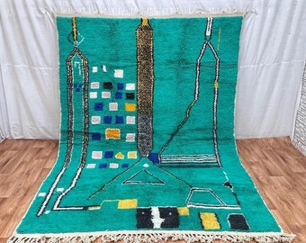 May include: A hand-woven Moroccan rug with a geometric design in shades of teal, white, black, yellow, and pink. The rug has a fringe border.