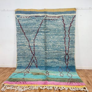 May include: A large, rectangular rug with a textured, light blue background and geometric maroon designs. The rug features a colorful border with pink, yellow, and green stripes, and white fringe along the bottom edge. The rug is displayed against a white wall.
