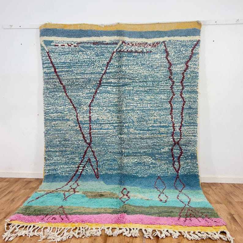 May include: A large, rectangular rug with a textured, light blue background and geometric maroon designs. The rug features a colorful border with pink, yellow, and green stripes, and white fringe along the bottom edge. The rug is displayed against a white wall.