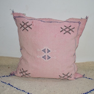 Puede incluir: Una almohada rosa con un patrón geométrico en hilo blanco y azul. La almohada tiene un borde de flecos y está hecha de una tela suave y tejida.